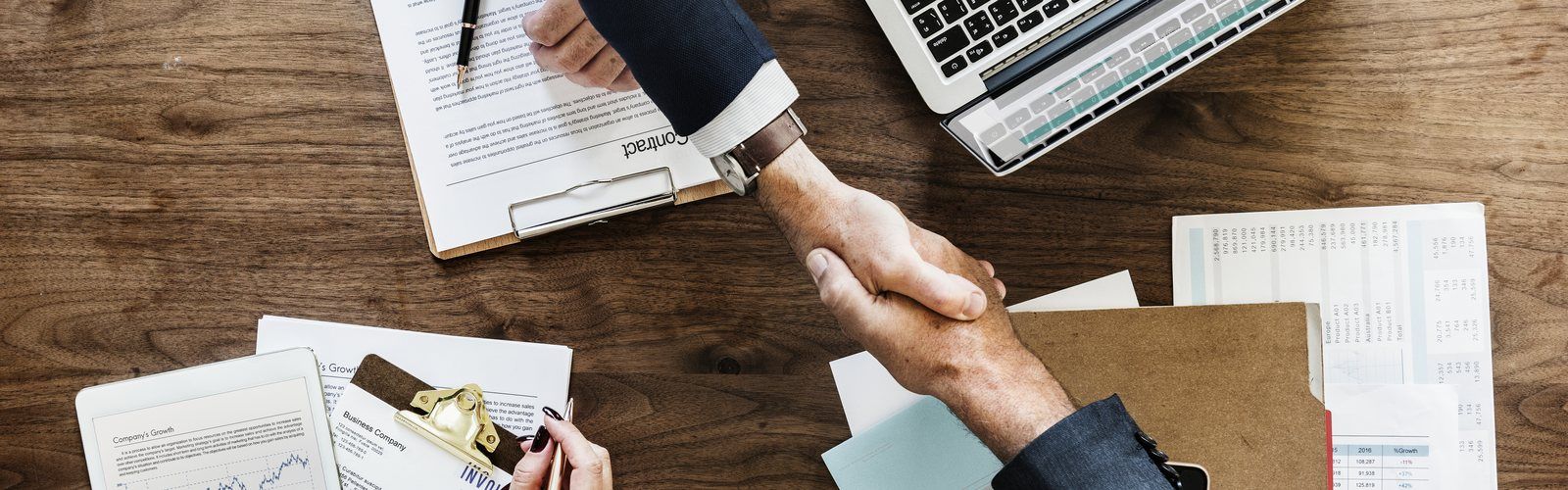 Two people shaking hands over a desk with a contract and financial documents